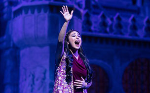 A performer in a purple and pink costume singing on stage with one arm raised in front of a blue castle set