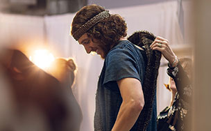 An actor in a headband and tunic being helped into a patterned robe backstage under stage lighting