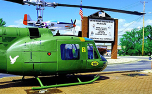 A green military helicopter displayed beside the Veterans Memorial Museum sign, with a propeller aircraft and an American flag behind