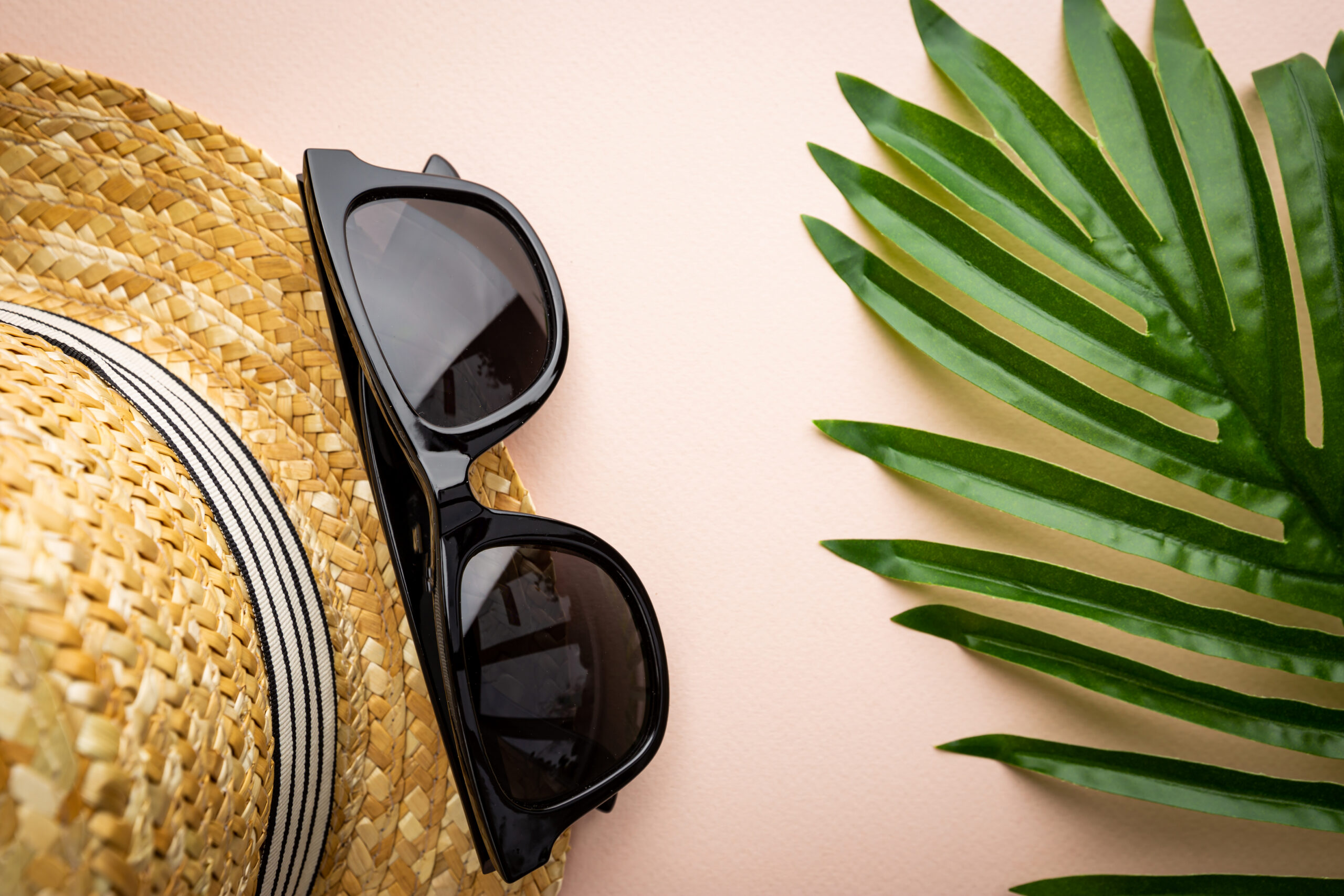 A straw sun hat and a pair of black sunglasses laid flat beside a green palm leaf on a pale pink background